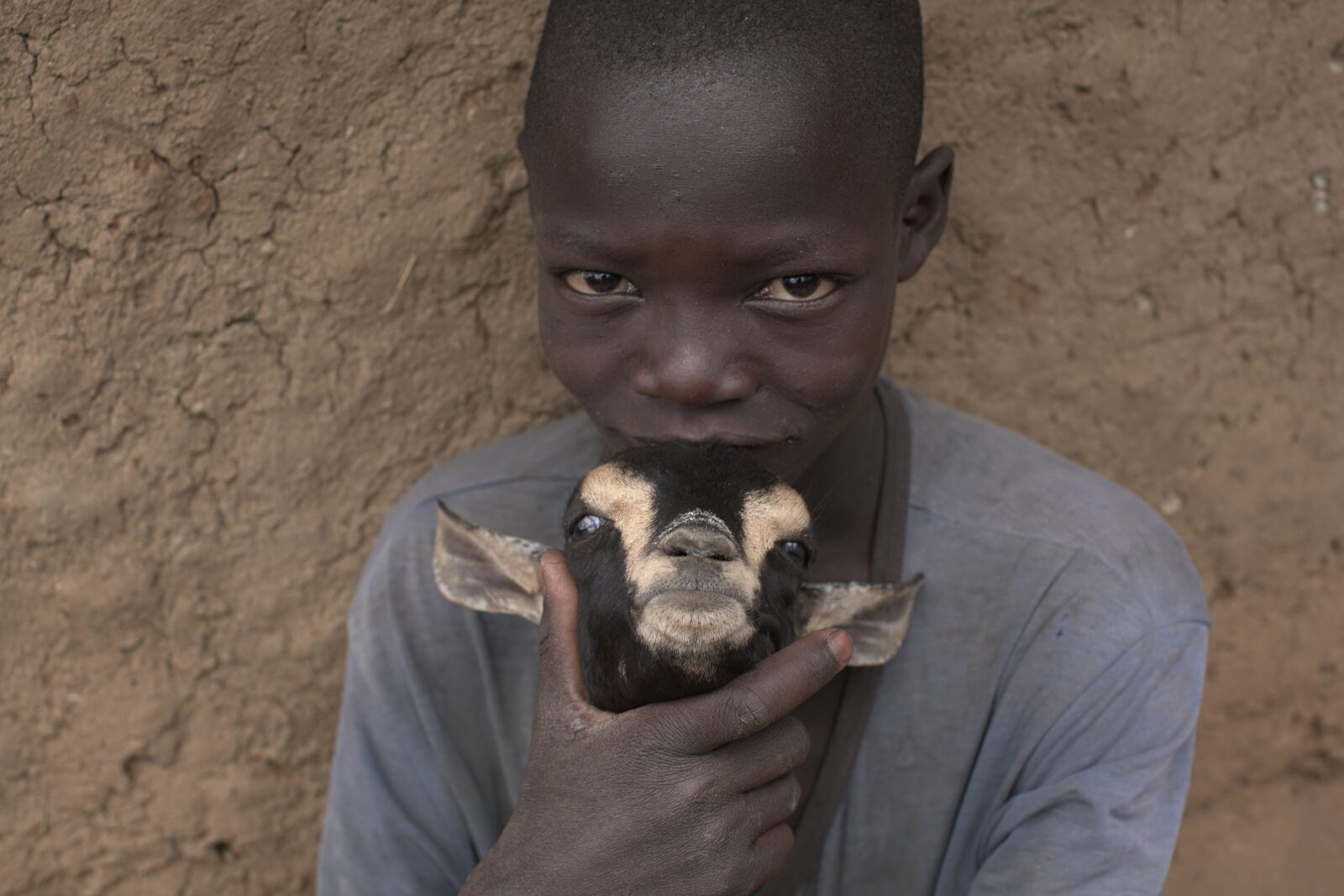 Un jeune garçon tient une chèvre contre son visage, devant un mur de terre, en Ouganda rural