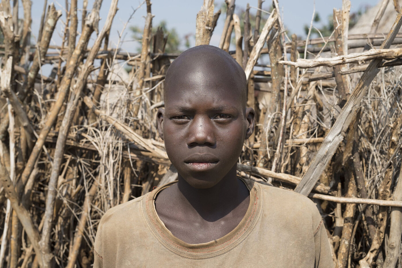 Lochap, the second young person followed in Karamoja Rising, standing before a woven stick fence