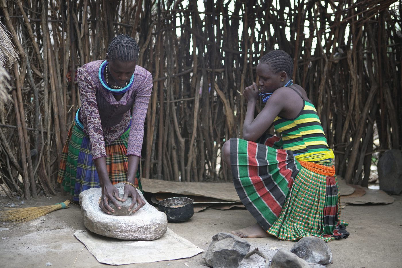 Sylvia and Norah grinding grain on a stone outside a woven stick fence