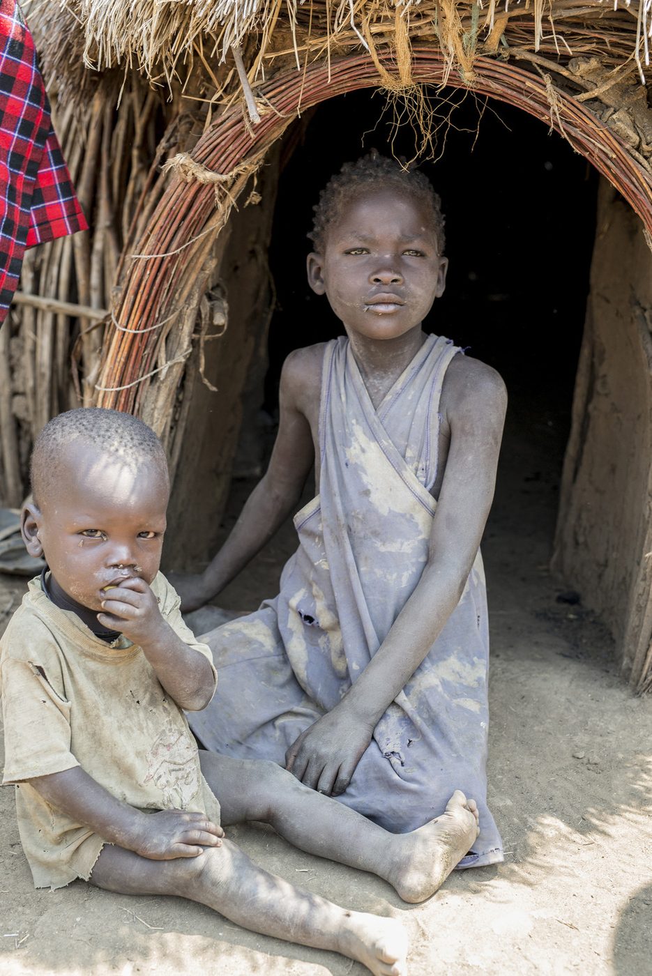 Two young children in torn clothing sitting in the doorway of a stick-and-mud home